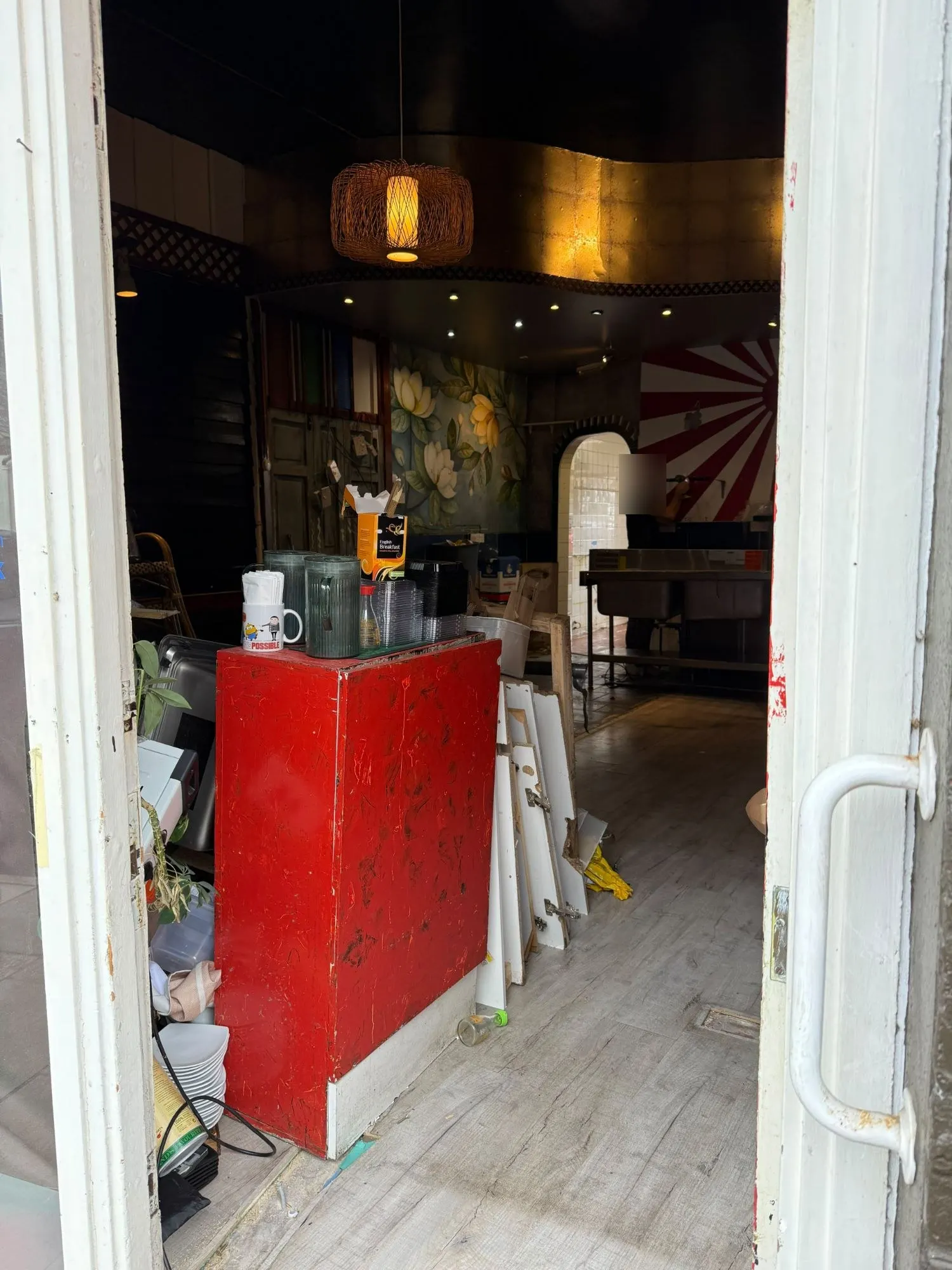 Looking through Tomi Sushi Bar's open door: a red counter piled with jugs and takeaway tubs, panels stacked against it, and the rising-sun mural and kitchen beyond