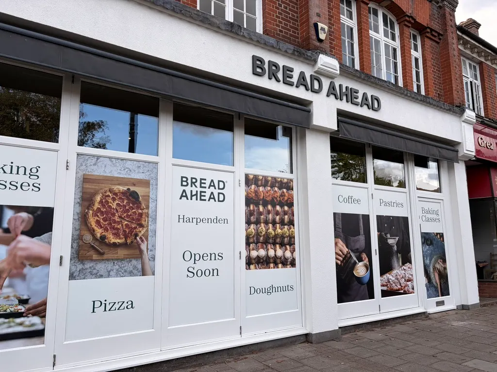 Bread Ahead's white shopfront on Harpenden High Street, with window graphics for pizza, doughnuts, coffee, pastries and baking classes and an 'Opens Soon' sign