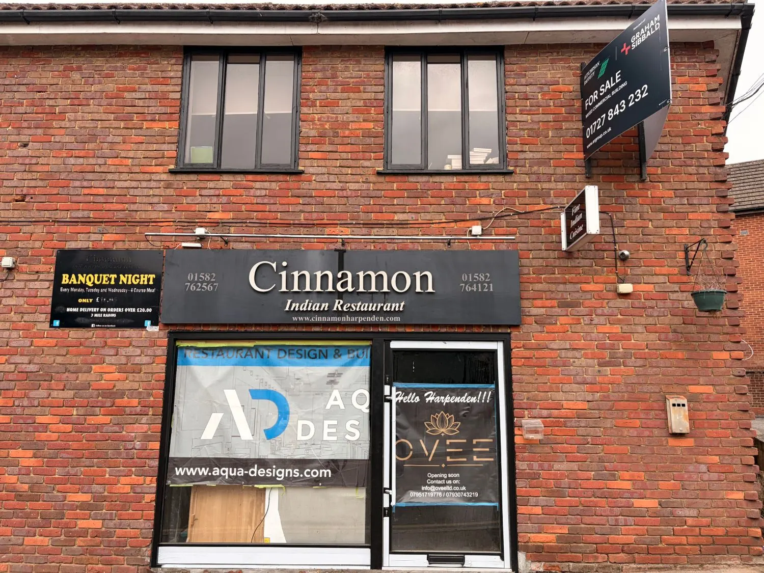 A brick building in Thompsons Close with the Cinnamon Indian Restaurant sign still above the shopfront, a restaurant fitters' banner in the window and an OVEE poster taped to the glass door