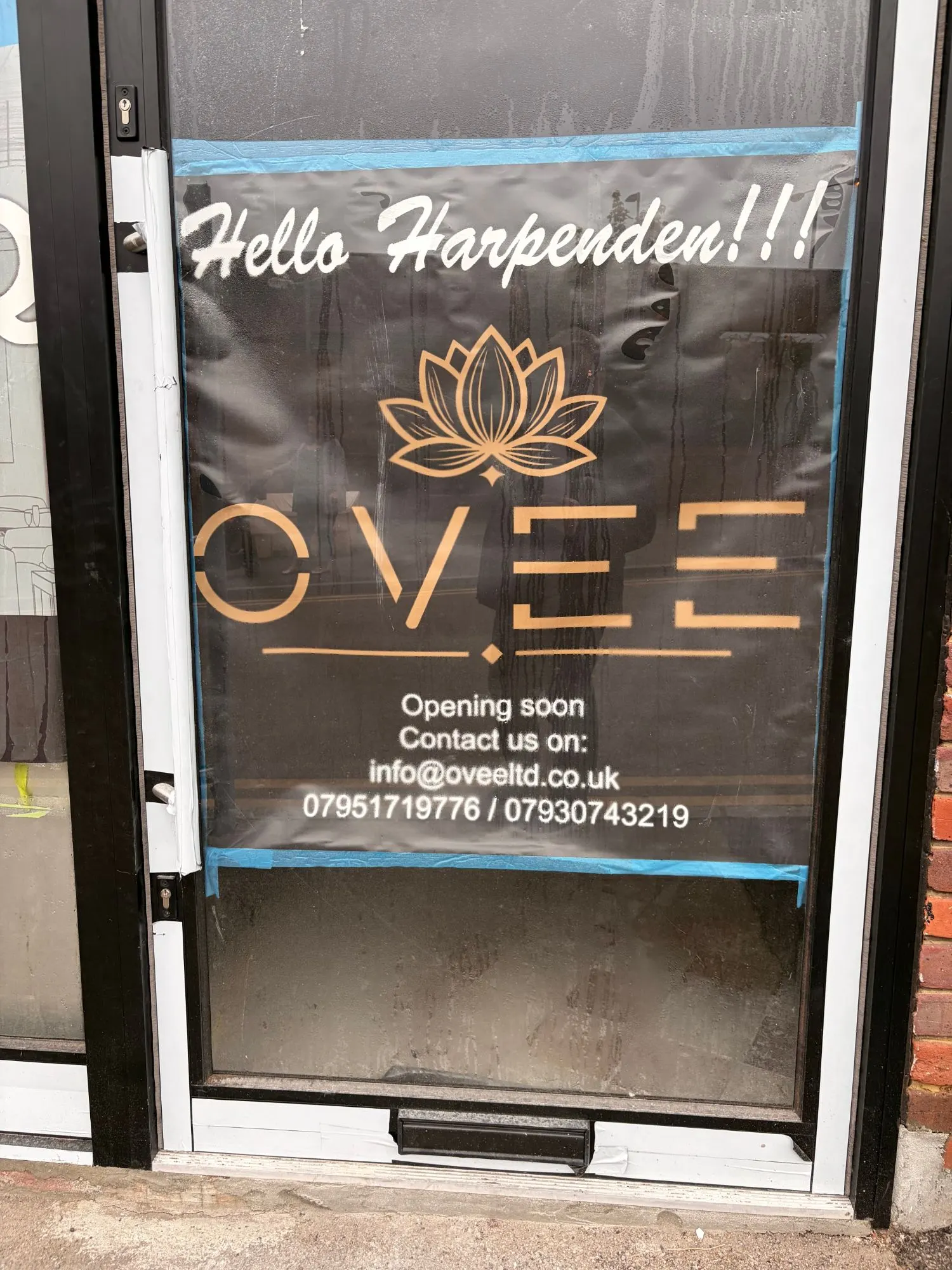 OVEE's poster on the restaurant's glass door, reading "Hello Harpenden!!!" above a gold lotus logo and the words "Opening soon"